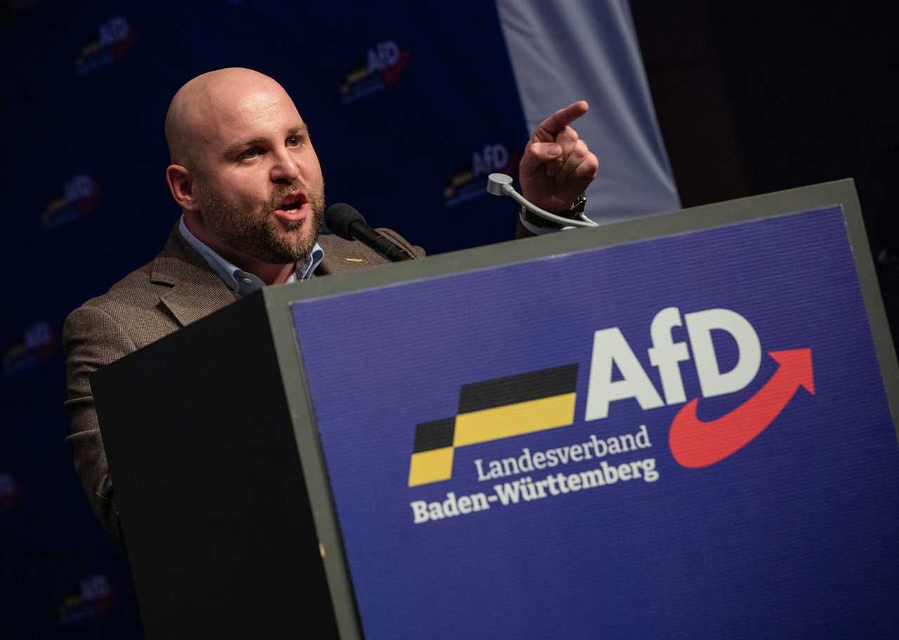 A white, bald middle aged man points his finger and stands at a podium that has the words 'AfD' and German writing on it.