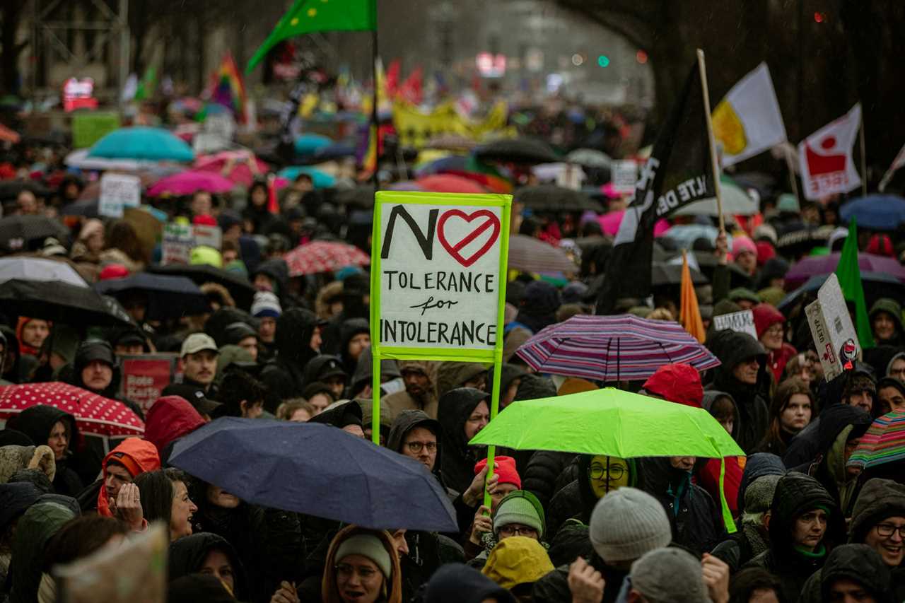 A large crowd of people stand close together with umbrellas and hold signs. One of them says 'No tolerance for intolerance.'